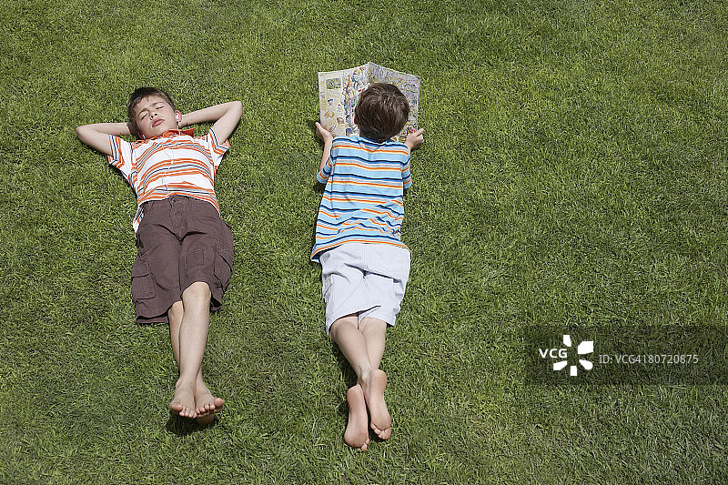 两个男孩(6-11岁)躺在草地上，一个在读高架读物图片素材