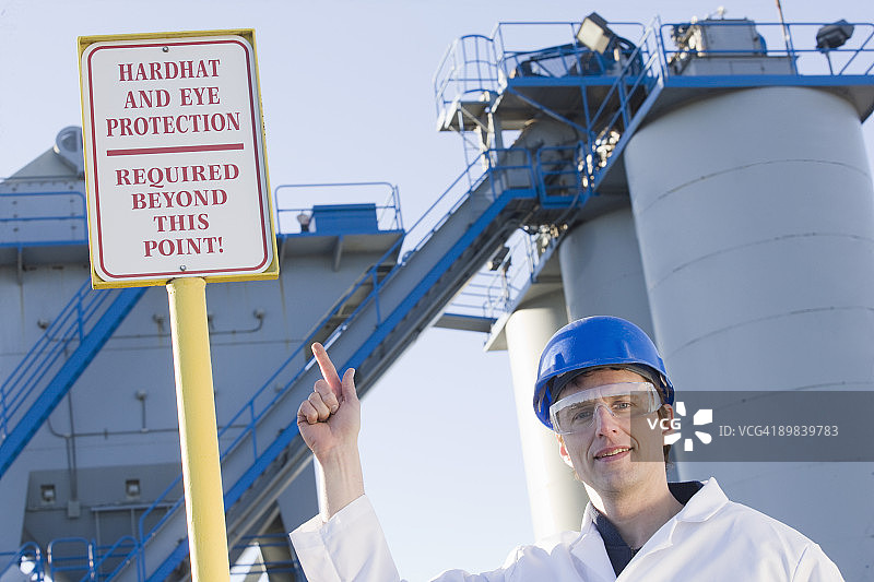 在一家沥青工厂，实验室工作人员指着一个警告标志，上面写着“过此点需要戴安全帽和保护眼睛”图片素材