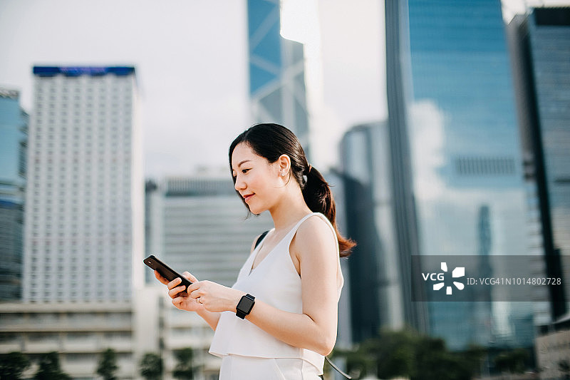 在城市里，微笑的年轻女商人用智能手机保持联系，背景是商业摩天大楼图片素材