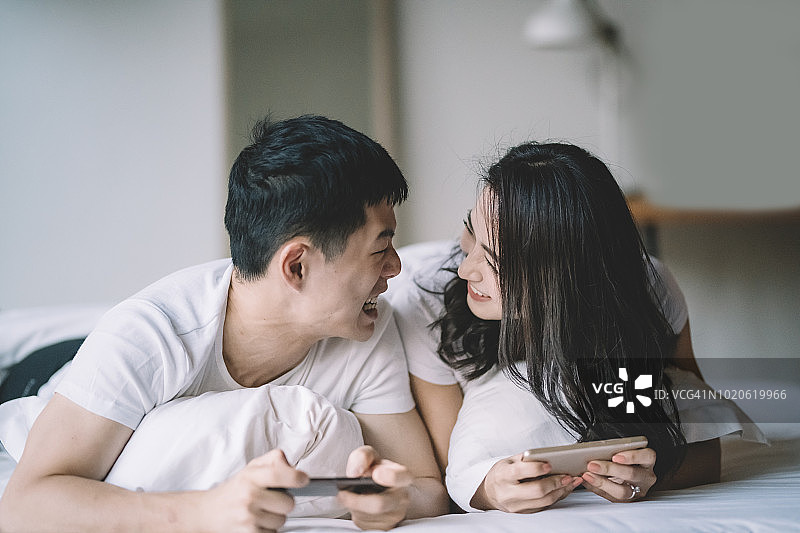 一对亚洲华人夫妇躺在床上玩手机游戏，早上玩得很开心图片素材
