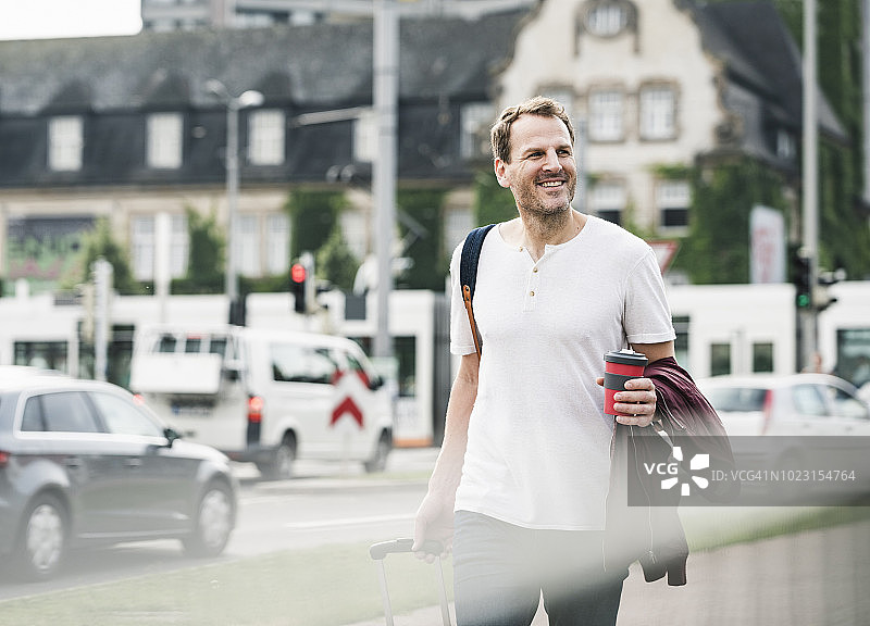 带着滚动行李箱和外卖咖啡走在城市里的微笑男人图片素材
