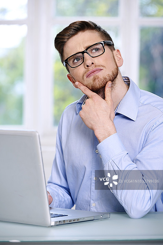 男子身穿蓝色衬衫，戴着眼镜，蓄着胡须，神情严肃，窗外的景物模糊不清，坐在电脑前的笔记本电脑前;向上看，两根手指放在下巴上，向上看。图片素材