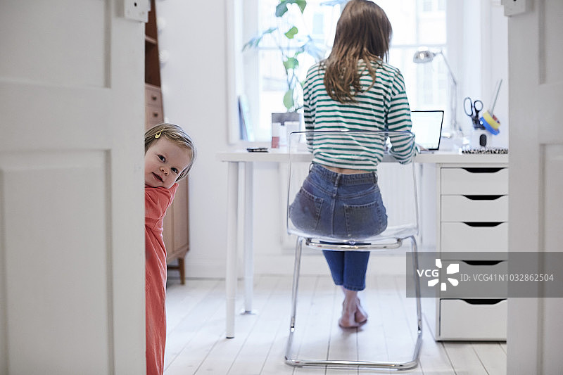 女孩站在门后，而母亲在后台工作的笔记本电脑在家里图片素材