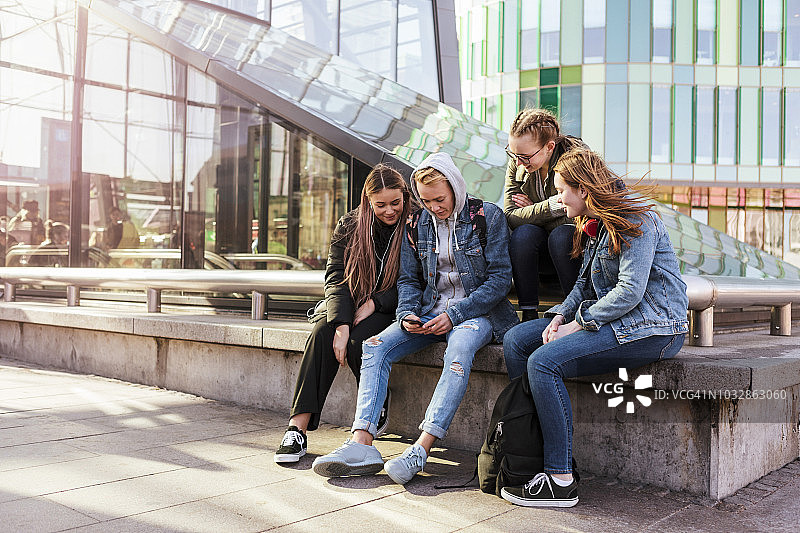 十几岁的女孩坐在城市的栏杆旁看朋友用手机图片素材