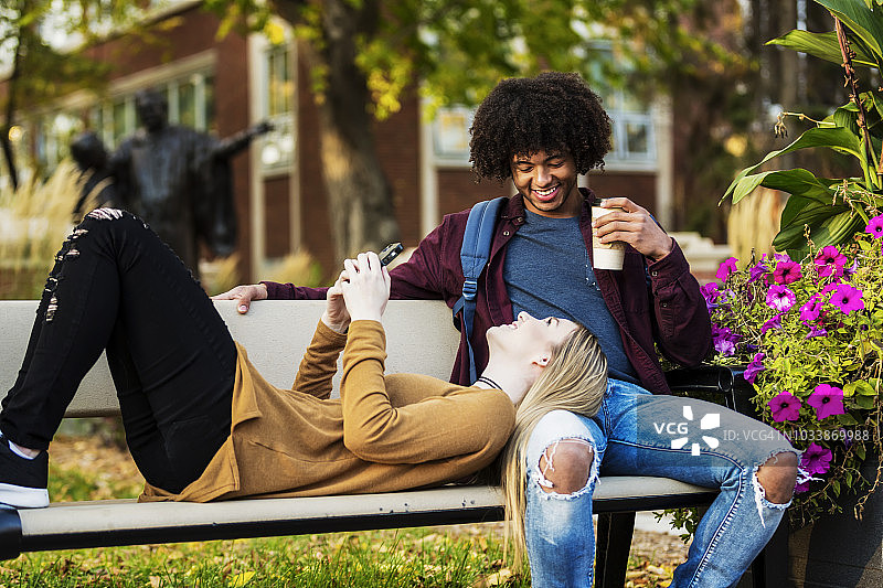 一对年轻的约会情侣在大学校园的长椅上放松，女人在看她的智能手机，把头放在她男友的腿上图片素材