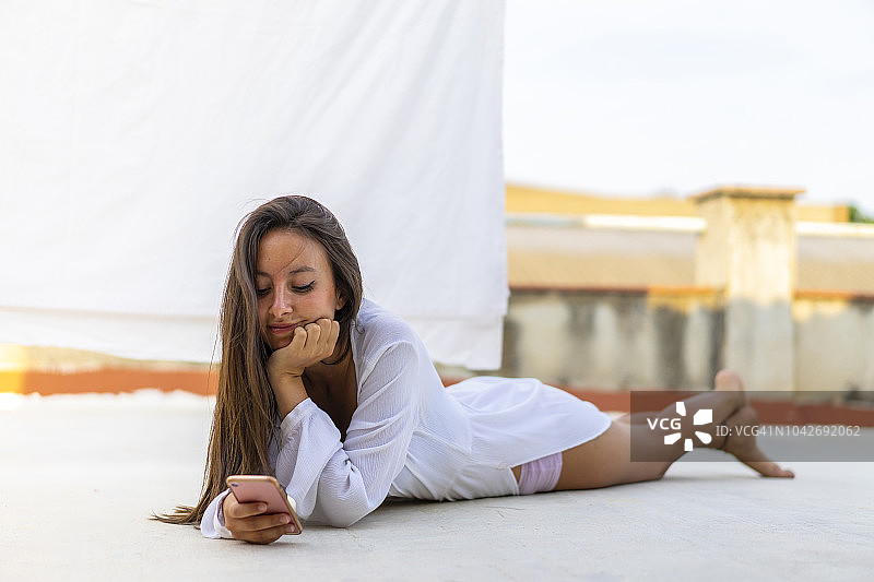 微笑的年轻女子躺在屋顶阳台上看手机的肖像图片素材