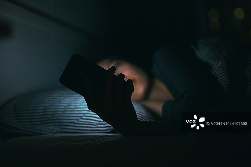 一名年轻女子晚上躺在床上休息时用智能手机发短信图片素材