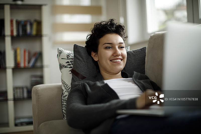 微笑的年轻女子使用笔记本电脑，而放松在家里的沙发上图片素材