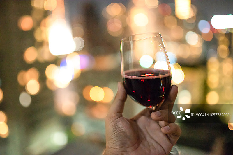 一个男人的手拿着一个红酒杯，背景是城市灯光的散景图片素材