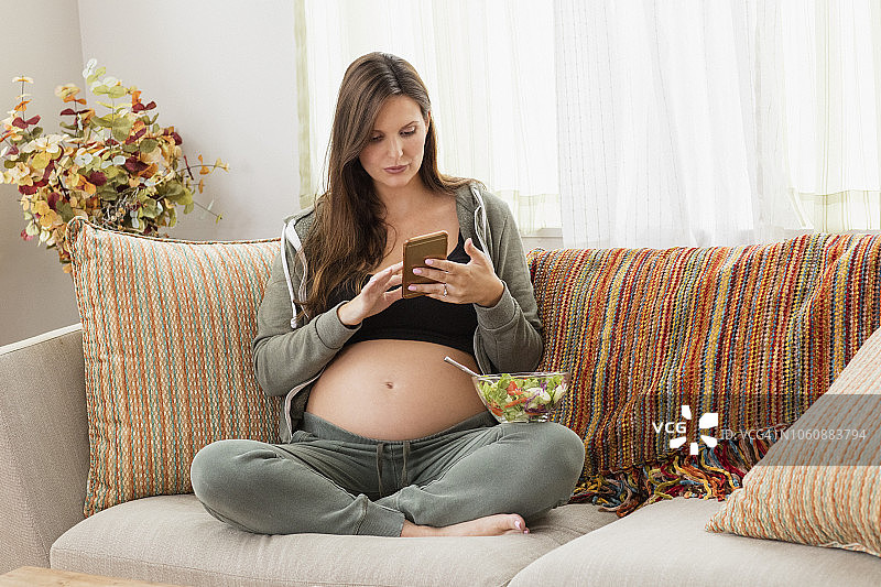年轻孕妇坐在沙发上吃沙拉，发短信图片素材