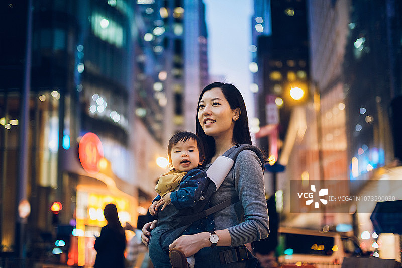 年轻的母亲和女婴在繁忙的街道上探索，在灯火通明的建筑物和繁忙的交通在城市的夜晚图片素材