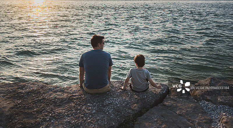 父亲和儿子坐在海边的岩石在日落的背影图片素材