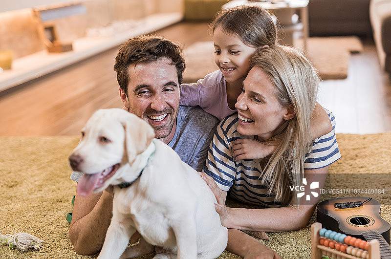 一个年轻幸福的家庭和一只小狗在客厅的地毯上休息。图片素材