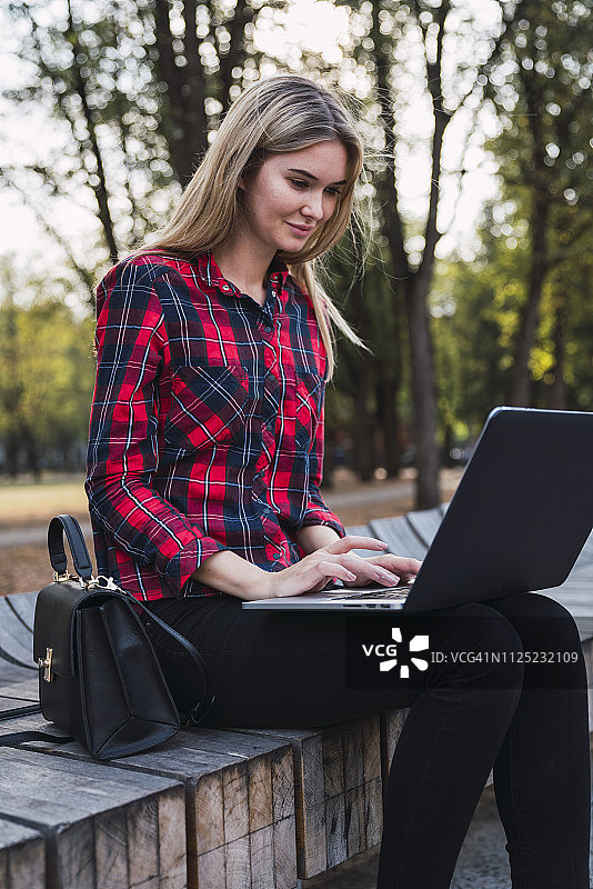 时尚的年轻女子坐在户外长椅上用笔记本电脑工作图片素材