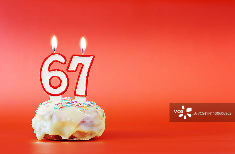 六十七岁生日。纸杯蛋糕，上面有燃烧着的白色蜡烛，数字是67。鲜艳的红色背景与复制空间图片素材