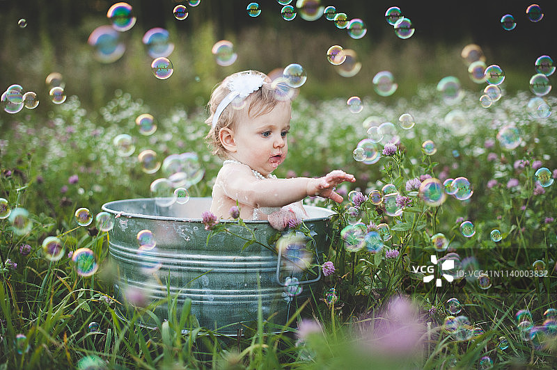 在草地上的锡浴缸里的女婴伸手去抓浮着的泡泡图片素材