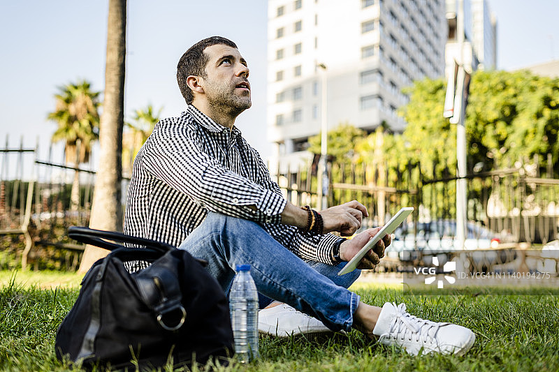 一名男子坐在城市公园的草地上，用数码平板电脑抬头看图片素材
