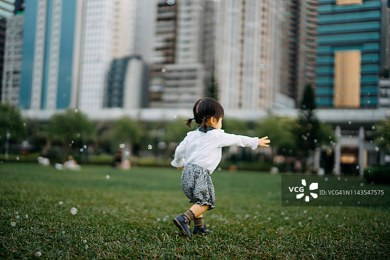 可爱的蹒跚学步的小女孩在城市公园草坪上的绿草上玩肥皂泡图片素材