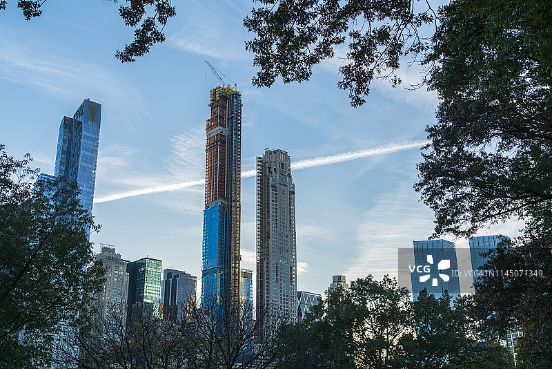 2018年11月4日，美国纽约曼哈顿中城摩天大楼在中央公园的树林中，飞机在秋日的蓝天上勾勒出凝结的痕迹。图片素材
