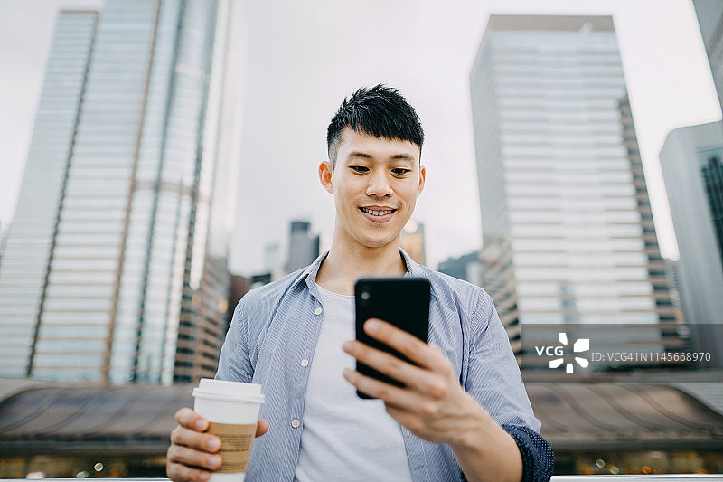 聪明的年轻亚洲男子享受咖啡和使用智能手机对着现代城市的天际线城市露台图片素材