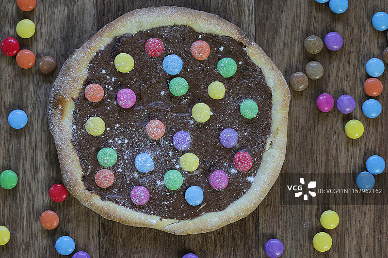 图片自制的巧克力披萨与五彩糖果糖果和糖霜糖，烤披萨皮充满榛子巧克力涂抹儿童，顶部是脆糖涂彩虹smarties巧克力糖果，吃布丁甜点图片素材
