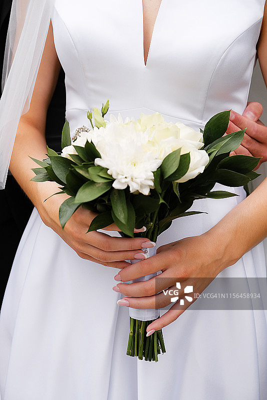 新娘身着优雅的婚纱，手捧一束美丽的白玫瑰、菊花和绿叶。拥抱新婚夫妇，新娘和新郎的手特写，户外。图片素材