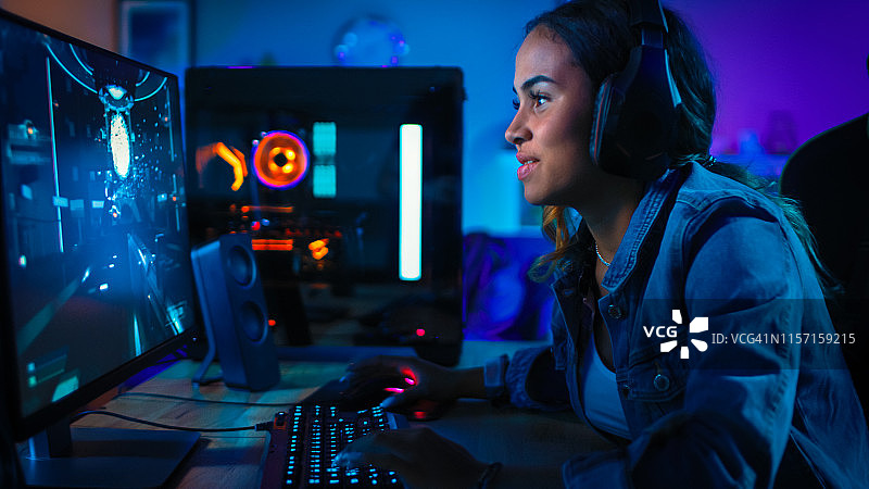 戴着耳机的漂亮和兴奋的黑人玩家女孩正在她的电脑上玩第一人称射击在线视频游戏。房间和电脑都有彩色霓虹灯。温馨的家庭之夜。图片素材