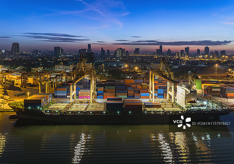 黄昏时分，以城市景观为背景的码头工作吊车桥将集装箱装载到集装箱船上。物流，进出口，海运或运输。图片素材