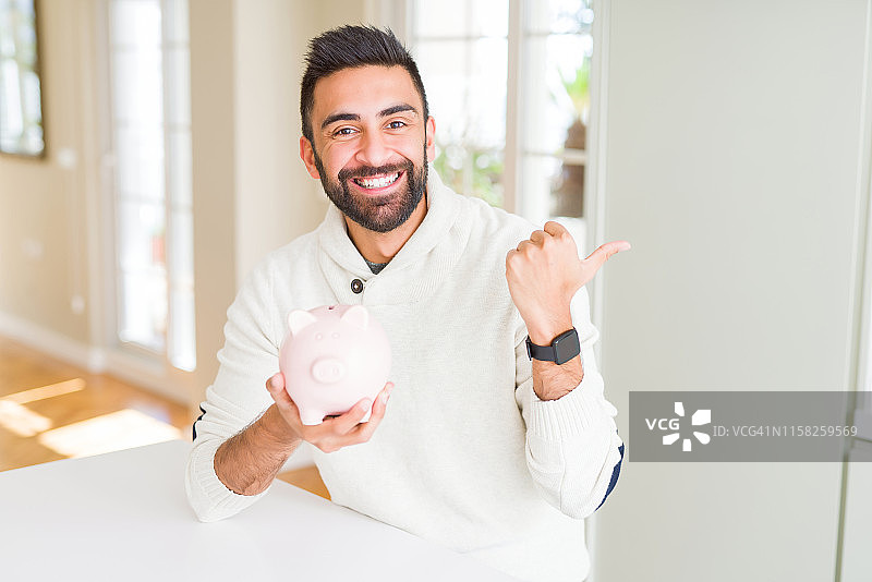 英俊的拉美裔男子拿着储蓄罐指向和显示与拇指向上到一边与快乐的脸微笑图片素材