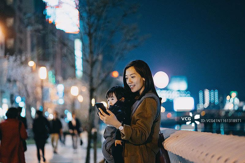 年轻的亚洲母亲抱着可爱的小女儿在城市的人行道上使用智能手机图片素材