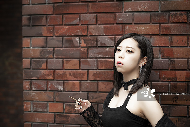 年轻美丽的亚洲女人吸烟。她靠在砖墙上。图片素材