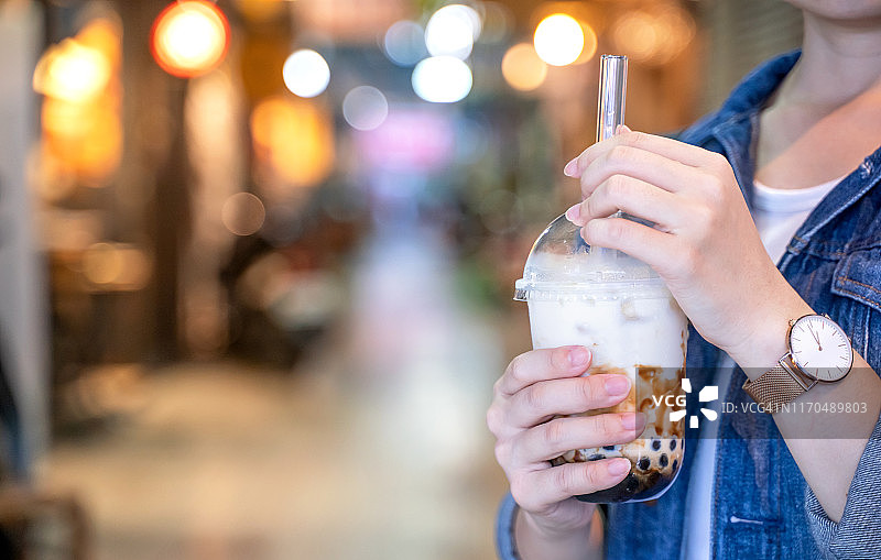 台湾夜市上，穿着牛仔夹克的小女孩正用玻璃吸管喝着红糖味的木薯珍珠奶茶。图片素材