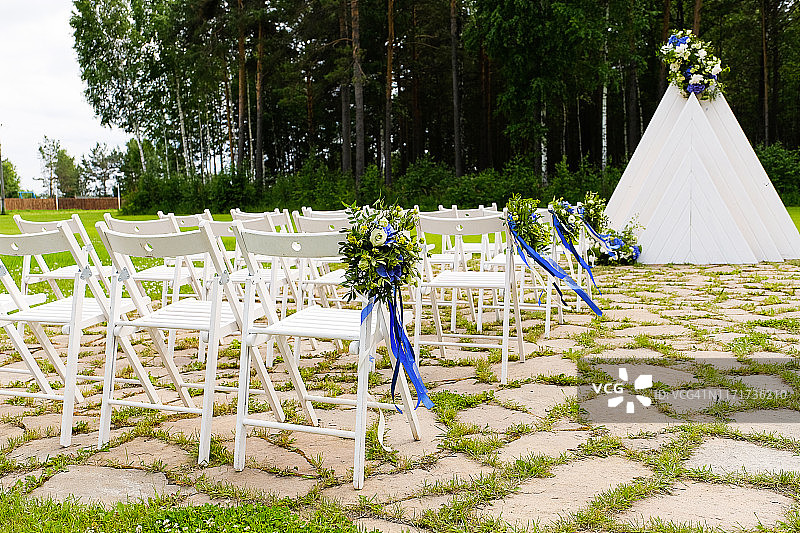 白色的木椅上装饰着花束和明亮的缎带，这是户外松树林里的婚礼装饰。图片素材