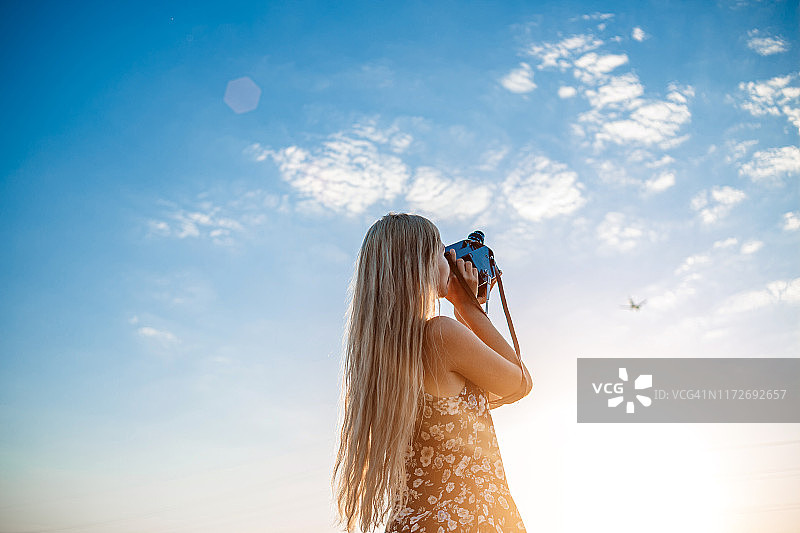 在葡萄地里，一个身穿印花连衣裙的金发女孩用老式摄像机拍摄了一架飞机起飞的视频图片素材