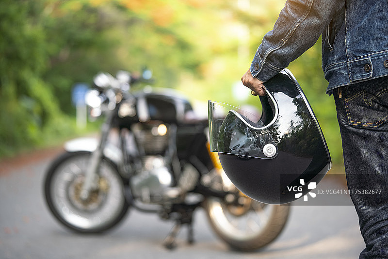 英俊的男子在牛仔裤是拿着一个头盔和老式摩托车模糊的背景图片素材
