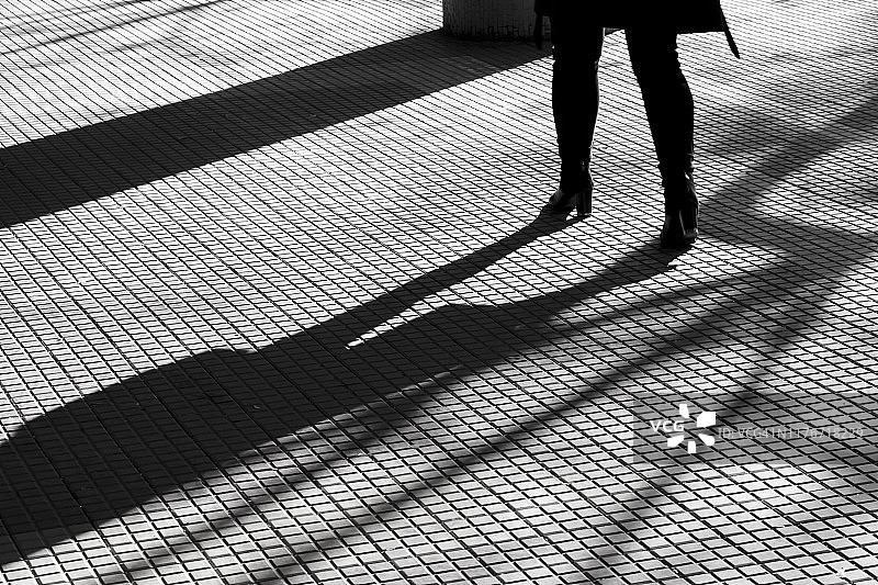 一个女人穿着高跟鞋独自走在铺着瓷砖的人行道上，腿的模糊剪影图片素材