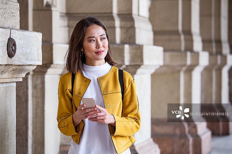 美丽的亚洲女人使用一个应用程序发送短信在她的智能手机设备。快乐的年轻亚洲女人拿着智能手机站在街上图片素材