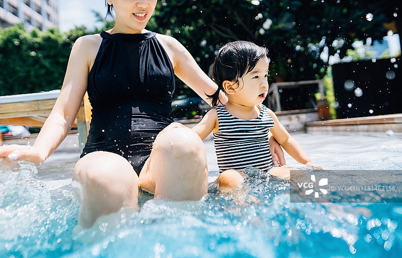 可爱的小亚洲学步女孩有乐趣在游泳池和她的母亲。他们挨个坐在泳池边溅起水花图片素材