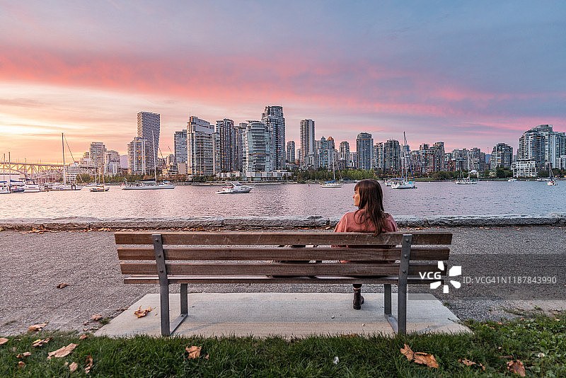 一个女人坐在长椅上看着温哥华的天际线，从岛上公园散步。加拿大不列颠哥伦比亚省图片素材