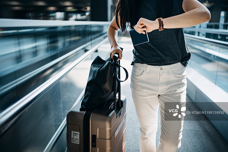 年轻的亚洲女子拿着行李箱和护照走在机场大厅时，手表上显示时间图片素材