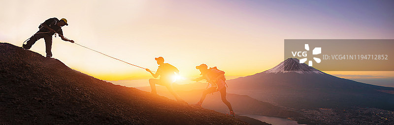 一群背着背包的旅行者越过日出的背景。年轻的亚洲三名徒步旅行者正在攀登富士山附近的山峰。人们互相帮助往上爬。伸出援手。帮助和团队合作的理念图片素材