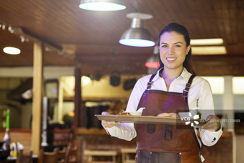 一名拉丁女服务员，黑头发，大约29岁，穿着白色衬衫制服，系着皮围裙，手里拿着一个托盘，上面是哥伦比亚最美味的菜肴，是在城里最好的餐馆里做的图片素材