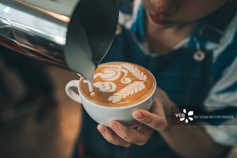 专业的咖啡师将蒸牛奶倒入咖啡杯中，制作美丽的罗塞塔拉花图案。在咖啡店喝一杯拿铁咖啡图片素材