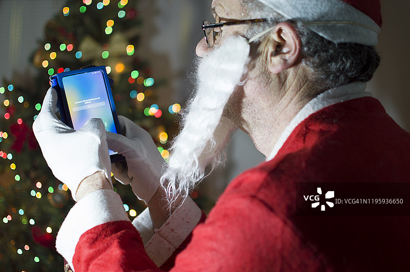 一名装扮成圣诞老人的男子在圣诞树前使用智能手机图片素材