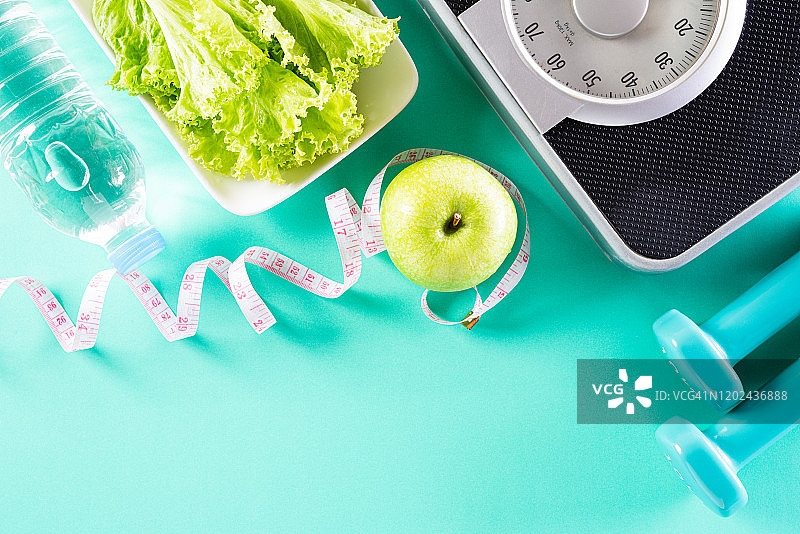 健康的生活方式、饮食和运动理念。俯视图运动员的设备重量秤测量带蓝色哑铃，运动水瓶，水果和蔬菜在明亮的绿色柔和的背景。图片素材