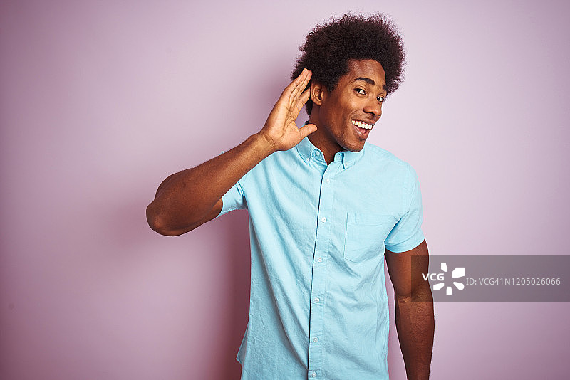 一名年轻的美国男子，留着非洲式发型，穿着蓝色衬衫，站在孤立的粉红色背景前微笑，捂着耳朵听谣言或流言蜚语。耳聋的概念。图片素材