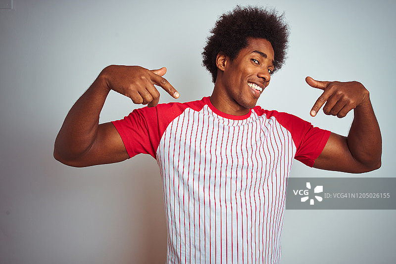 非洲裔美国人与非洲式头发穿红色条纹t恤在孤立的白色背景看起来自信与微笑，用手指指着自己骄傲和幸福。图片素材