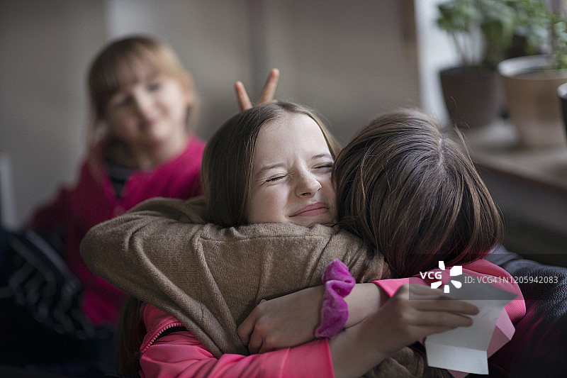 十几岁的女孩在收到礼品券后拥抱姐姐以表示感激，而妹妹在背景中做了有趣的兔子耳朵手势图片素材