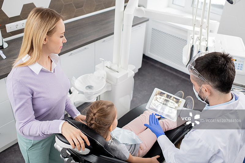 一个年轻女子站在她的小女儿旁边，坐在扶手椅上看牙医图片素材