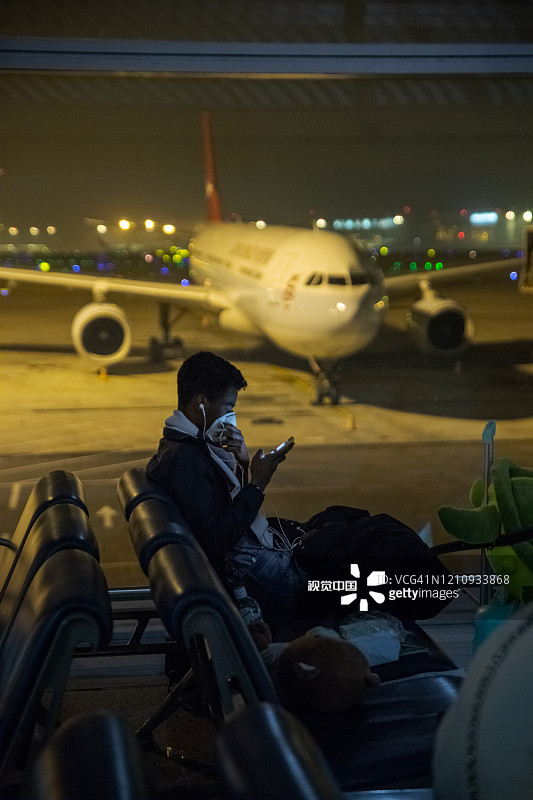 2019冠状病毒(covid-19)疫情爆发，北京国际机场，游客(深色皮肤)戴着卫生口罩看手机，因为病毒的危险图片素材